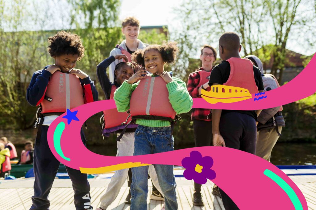 A group of children wearing red life jackets smile and pose on a wooden dock near a body of water, with two adult supervisors in the background. The children look happy and excited, possibly preparing for a water-based activity. The image features playful pink illustrated swirls overlaid with cartoon graphics of a boat, flower, and star, adding a fun, vibrant touch. Trees and buildings are visible in the sunny background.
